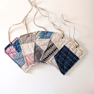 May include: A collection of six quilted fabric gift tags in various colors and patterns. Each tag is a unique patchwork design with a neutral-colored string for hanging. The tags are arranged in a fan shape against a white background.