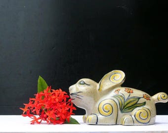 Könnte beinhalten: Eine Keramik-Hasenfigur mit gelben und grünen floralen Akzenten und Wirbeldetails. Leuchtend rote Blumen und ein grünes Blatt befinden sich neben dem Hasen. Der Hase steht auf einer weißen Oberfläche mit schwarzem Hintergrund.