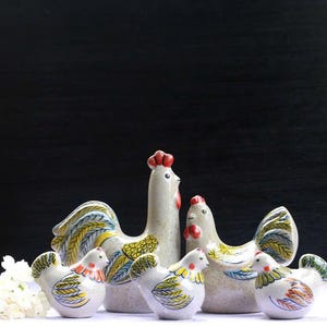 May include: A ceramic rooster family, featuring a tall rooster, hen, and three chicks. The figurines are white with colorful accents of yellow, blue, and red. The rooster has a red comb. The family is arranged on a white surface against a dark background.