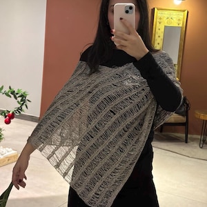 以下が含まれることがあります： フリンジ付きのシルバーグレーのオープン織りポンチョ。ポンチョは黒いトップスと黒いパンツの上に着用されています。オープン織りのデザインは、テクスチャード加工された軽量の外観を作り出しています。