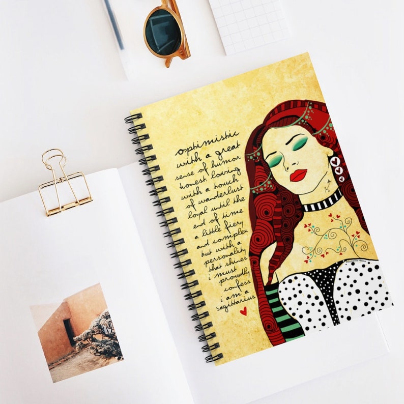 May include: A yellow notebook with a spiral binding features a colorful illustration of a woman with red hair and a black and white polka dot dress. The text on the notebook reads "optimistic with a great sense of humor honest loving with a touch of wanderlust loyal until the end of time a little fiery and complex but with a personality that shines i must proudly confess i am a Sagittarius".