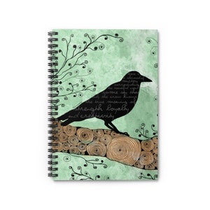 May include: Spiral-bound notebook featuring a raven silhouette on a log, set against a pale green backdrop. The design incorporates swirling branches and text about strength, loyalty, and creativity. The notebook's cover has a unique artistic design.