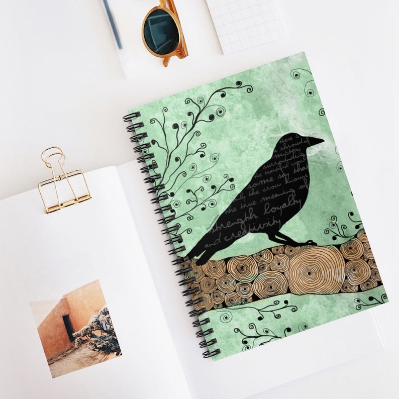 May include: A spiral-bound notebook with a raven design. The raven is black and sits on a log against a mint green backdrop. The design includes decorative swirls and text that reads "strength, loyalty, and creativity."