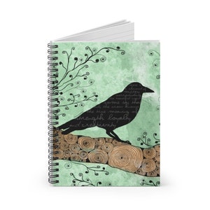 May include: Spiral-bound notebook with a black crow design on a light green background. The crow is perched on a log with a wood grain pattern. Text includes "strength, loyalty, and creativity". Decorative black swirls and text complete the design.