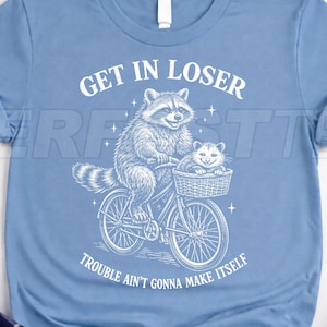 Può includere: T-shirt azzurra con un procione e un gattino in bicicletta, con la scritta "GET IN LOSER" e "TROUBLE AIN'T GONNA MAKE ITSELF". La grafica è bianca e le maniche sono arrotolate.