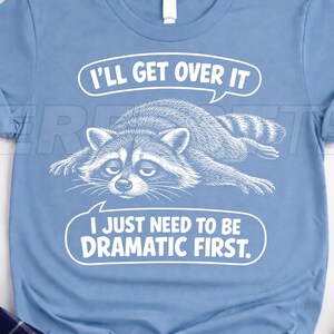 Op de afbeelding: Lichtblauw T-shirt met een afbeelding van een wasbeer en de tekst "I'll get over it, I just need to be dramatic first." De wasbeer ligt met een verdrietige uitdrukking.