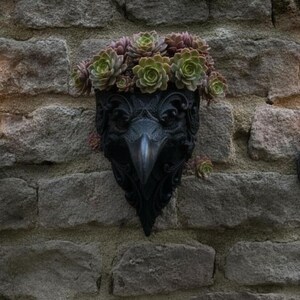 Könnte beinhalten: Ein schwarzer Wandpflanzkübel in Form eines Vogelkopfes mit detaillierten Schnörkeln, gefüllt mit grünen und violetten Sukkulenten, flankiert von zwei schwarzen Wandleuchtern aus Metall, die jeweils eine brennende weiße Stumpenkerze halten.
