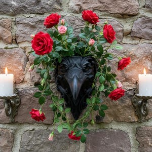 May include: A black wall-mounted raven head planter filled with vibrant red roses and green leaves, flanked by two antique-style metal wall sconces, each holding a single white pillar candle, all mounted against a rustic stone wall background.