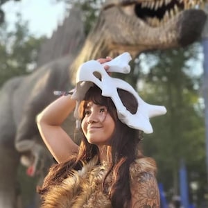 Puede incluir: Se sostiene un tocado blanco con forma de hueso, posiblemente un art&iacute;culo de disfraz o cosplay. La persona lleva un chaleco de piel sint&eacute;tica marr&oacute;n. Una gran estatua de dinosaurio est&aacute; en el fondo.