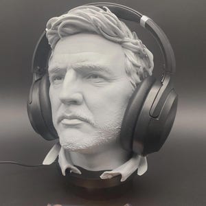 Peut inclure: Buste gris d'un homme portant un casque audio noir. Le buste présente des traits faciaux détaillés et est monté sur un socle noir. Le casque est positionné sur la tête, les écouteurs couvrant les oreilles. Un câble sort du casque.