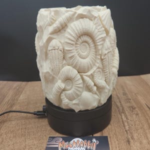 May include: A cream-colored, fossil-themed decorative container. The container is adorned with detailed relief carvings of various prehistoric marine fossils, including ammonites and trilobites. It sits on a black rotating base, with a business card in the foreground.