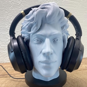 Peut inclure: Buste gris clair d'un homme avec un casque noir et des accents dorés. Le buste est posé sur une base circulaire noire. Le fond est un mur beige clair et une surface en bois. Le casque est posé sur la tête.