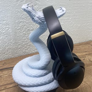 May include: A black over-ear headphone rests on a light blue snake-shaped headphone stand. The stand has a detailed, textured surface. The headphones have gold accents. The stand is on a wooden surface.