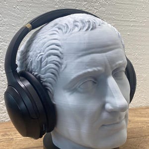 Puede incluir: Busto gris claro de una figura clásica con auriculares negros. El busto está sobre una base negra y los auriculares tienen detalles dorados. El fondo es una pared blanca texturizada y una superficie de madera.