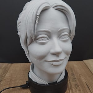 May include: A 3D-printed bust of a person with short hair and a slight smile. The bust is white and sits atop a black base with a power cord. A business card with a logo is visible.