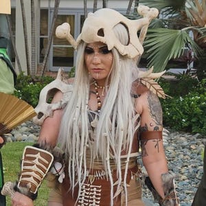 Puede incluir: Un disfraz de cosplay con un tocado de calavera con detalles de hueso, un corpi&ntilde;o de cuero marr&oacute;n y protectores de brazo. El disfraz incluye pelo largo y blanco y pintura facial tribal. El tema general es la fantas&iacute;a o el cosplay.
