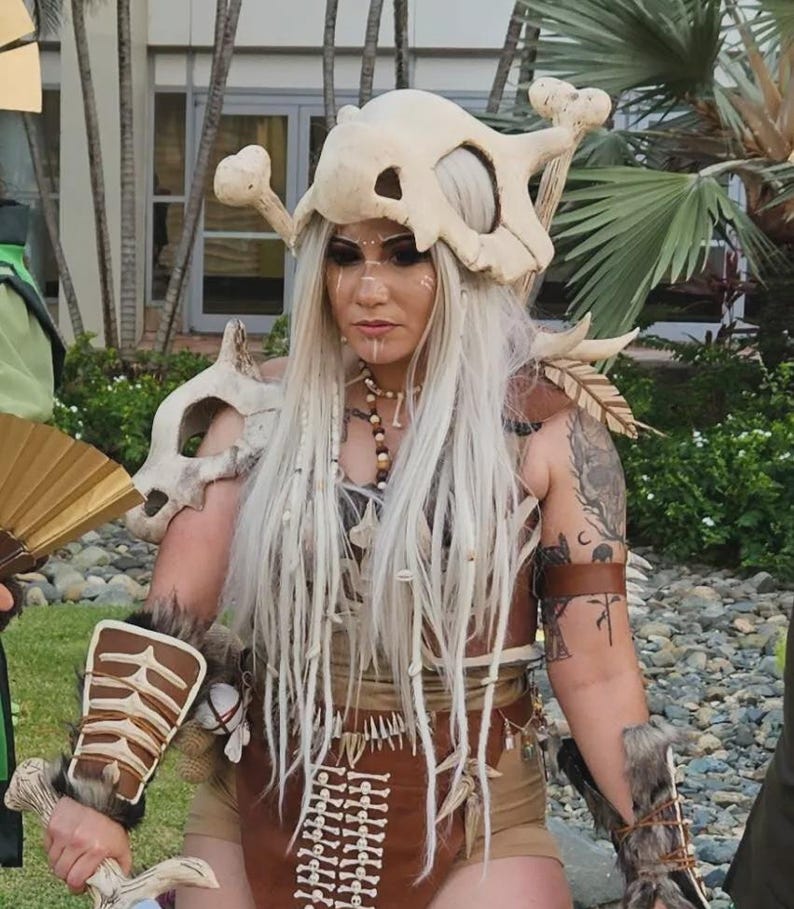 Puede incluir: Un disfraz de cosplay con un tocado de calavera con detalles de hueso, un corpi&ntilde;o de cuero marr&oacute;n y protectores de brazo. El disfraz incluye pelo largo y blanco y pintura facial tribal. El tema general es la fantas&iacute;a o el cosplay.