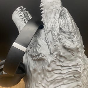 May include: A grey, 3D-printed sculpture of a roaring dinosaur head with sharp teeth. A black headband with a silver accent is placed over the dinosaur's head, creating a unique design. The sculpture has a textured surface.
