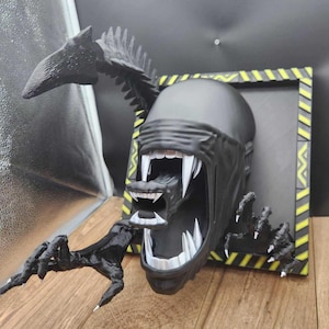 May include: A black wall-mounted sculpture of a Xenomorph head with an inner jaw extended, sharp white teeth, and clawed limbs emerging from a square frame featuring yellow and black hazard warning stripes along the outer border edges.