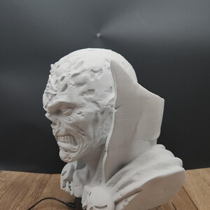 Puede incluir: Un busto blanco de un monstruo con un rostro detallado y texturizado, boca abierta y una capa con capucha. El busto se asienta sobre una base negra con un emblema de calavera. Una tarjeta de visita con el texto "MoshRabbit Trinkets" es visible.