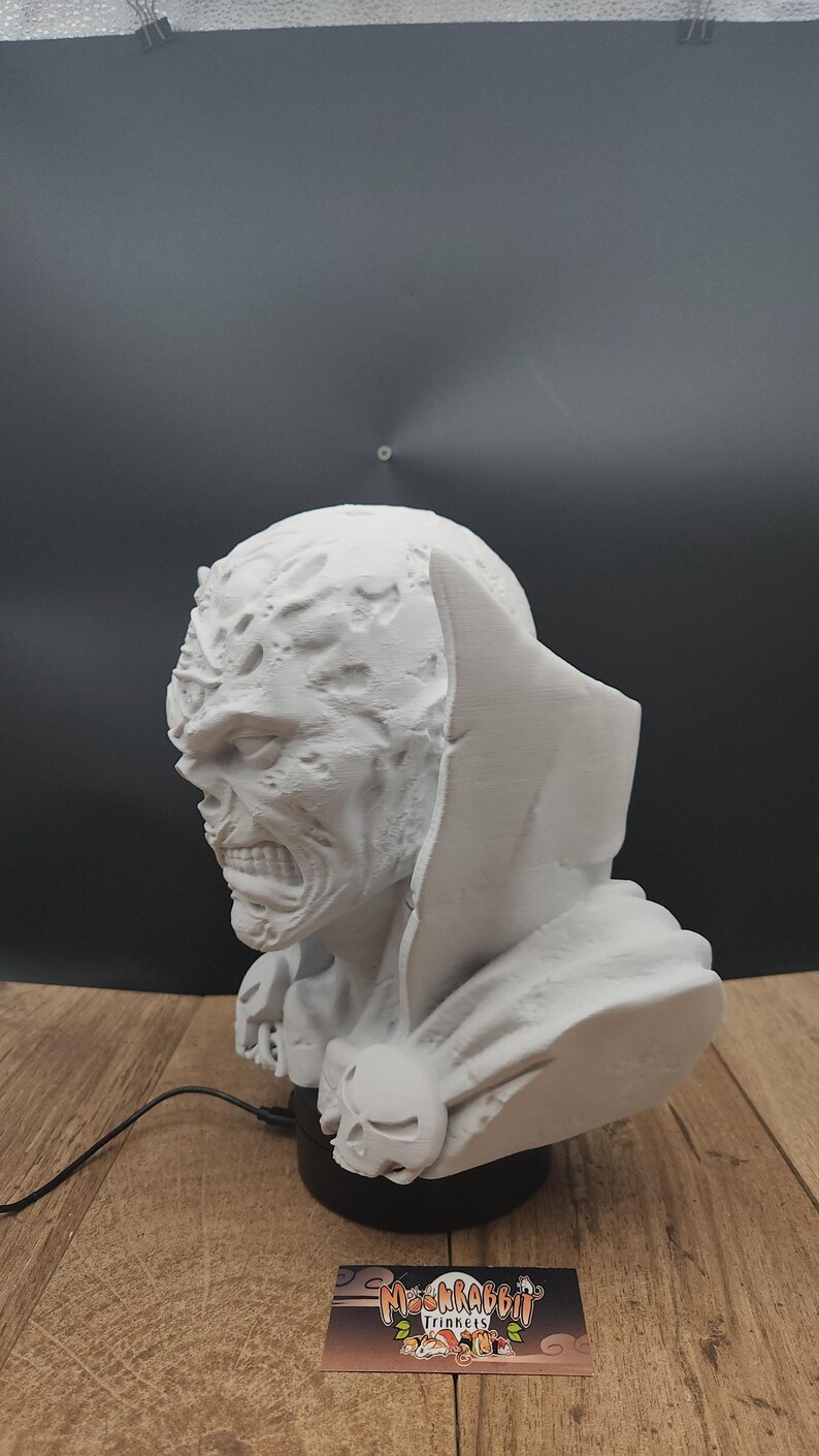 Puede incluir: Un busto blanco de un monstruo con un rostro detallado y texturizado, boca abierta y una capa con capucha. El busto se asienta sobre una base negra con un emblema de calavera. Una tarjeta de visita con el texto "MoshRabbit Trinkets" es visible.