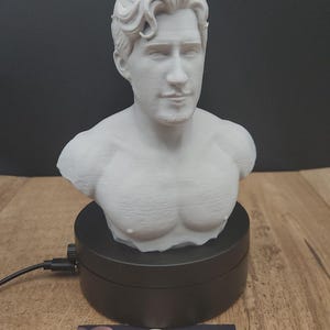 Pode incluir: Um busto branco de um homem com cabelo esculpido e peito musculoso. O busto é montado em um suporte de exibição giratório preto. O logotipo MoonRabbit Trinkets é visível na base.