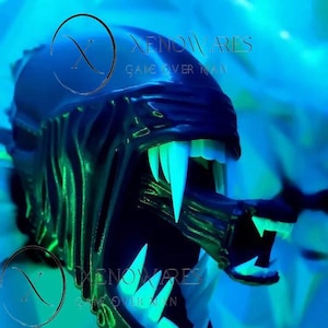 Puede incluir: Primer plano de una cabeza de Xenomorfo negra y verde con dientes blancos afilados y una boca interna prominente. La imagen tiene un brillo de neón azul y verde, con el texto "Xenowares Game Over Man" visible.