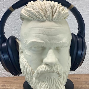 May include: A white bust of a bearded man wearing black over-ear headphones. The bust is on a black base with a charging cable. The headphones have gold accents.