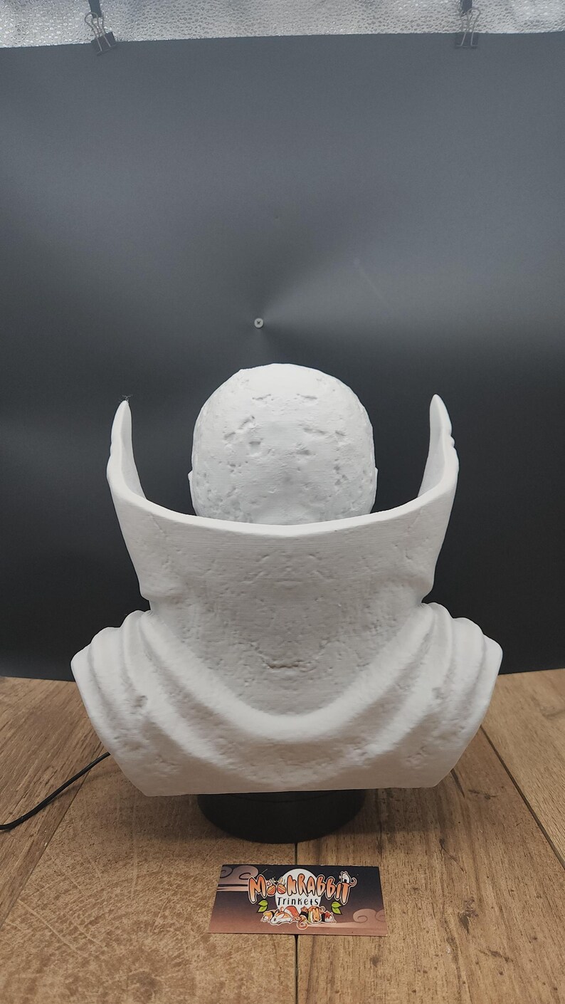 Puede incluir: Un busto blanco de un personaje con cabeza redondeada y cuello tipo capa. El busto est&aacute; sobre un fondo negro y sobre una base negra. Se ve una tarjeta de visita con el texto "Mr. Rabbit Trinkets".