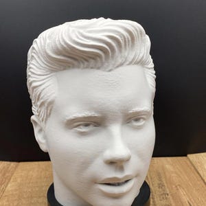 May include: A white bust of a man with styled hair. The sculpture is mounted on a black base. Two business cards with the text "Ms. Rabbit Trinkets" are in the foreground.