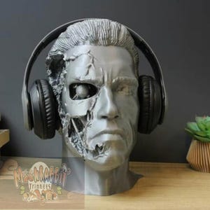 May include: A grey cyborg bust with headphones. The bust has a detailed, weathered appearance, with one side revealing mechanical components. Black headphones are placed over the head. The bust is on a wooden surface with books and plants in the background.