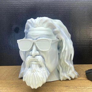 May include: A light blue bust of a bearded man wearing white sunglasses. The sculpture has long hair and a detailed beard. The bust is on a wooden surface with a dark background.