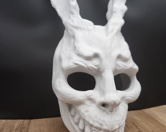 May include: A white, textured rabbit mask featuring long, upright ears, deep eye sockets, and a prominent, toothy grin. The mask displays intricate, organic-looking surface details and contours, with a small logo card labeled MoonRabbit Trinkets.