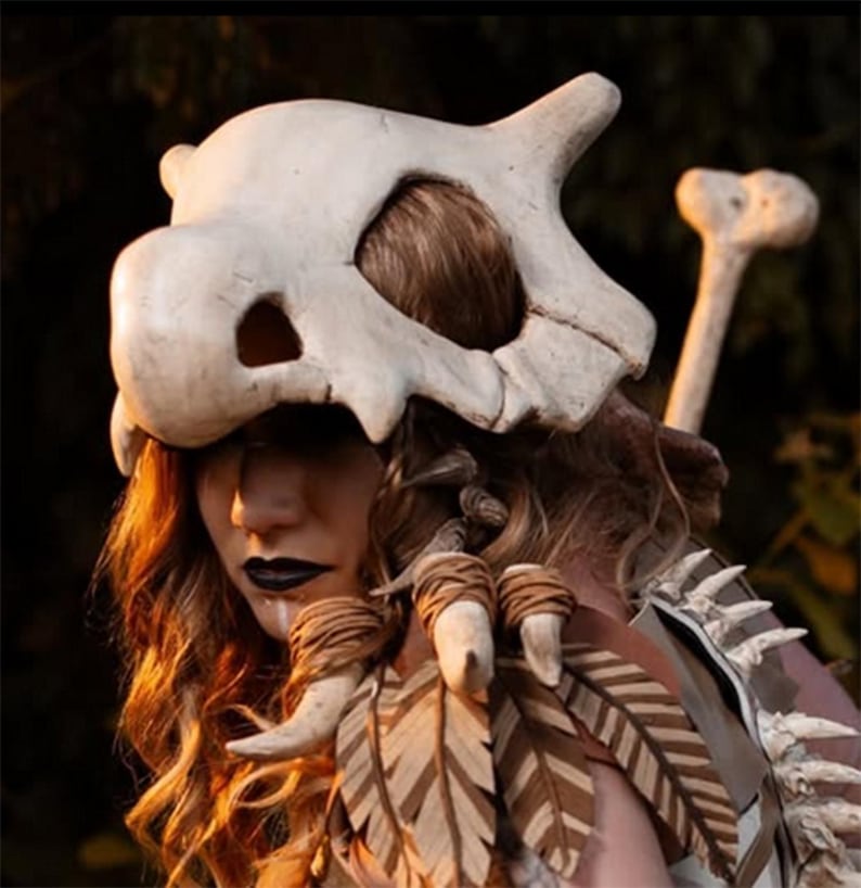 Puede incluir: Una m&aacute;scara de calavera de Cubone y un accesorio de hueso, parte de un disfraz de cosplay. La m&aacute;scara es blanquecina con cuencas oscuras y un borde inferior dentado. La hombrera presenta plumas de madera y detalles &oacute;seos.
