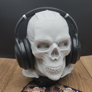 Puede incluir: Un soporte para auriculares con forma de calavera blanca con auriculares negros. La calavera tiene rasgos faciales detallados. Dos tarjetas de visita con el texto "Ms. Rabbit Trinkets" están en la base, sobre un fondo oscuro.