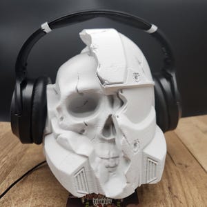 May include: A white skull-shaped headphone stand with a cracked design, paired with black headphones. The stand has a detailed, textured surface. The headphones are positioned over the skull, creating a unique display. A small card with text is at the base.