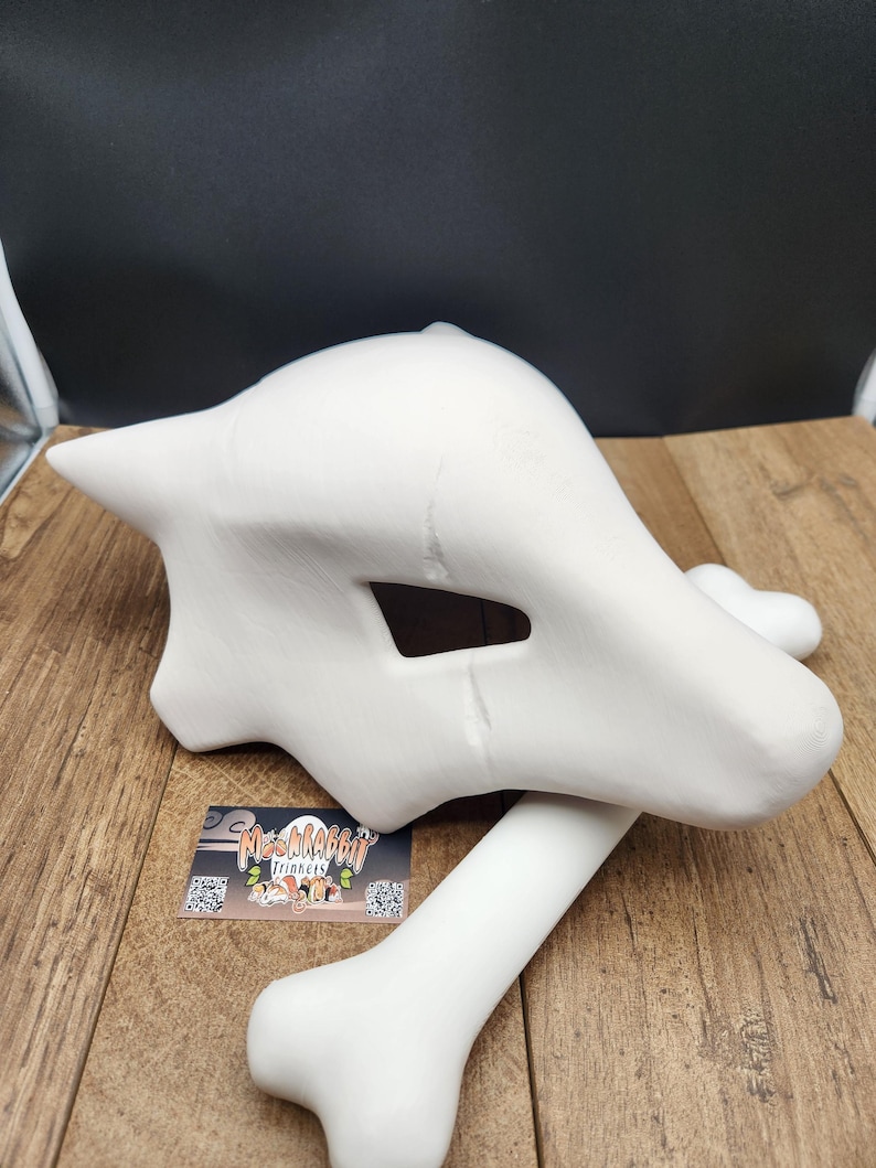 Puede incluir: Una m&aacute;scara de calavera Cubone blanca sin pintar y un accesorio de hueso. La m&aacute;scara presenta un dise&ntilde;o estilizado distintivo con aberturas para los ojos y la boca. El accesorio de hueso tiene una forma larga y curvada.