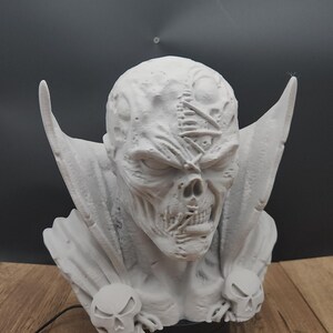 Puede incluir: Escultura detallada en gris claro de una cara esquel&eacute;tica con orejas grandes y puntiagudas y una capa. La escultura tiene una superficie texturizada y est&aacute; montada sobre una base negra. Dos detalles en forma de calavera en la parte delantera.