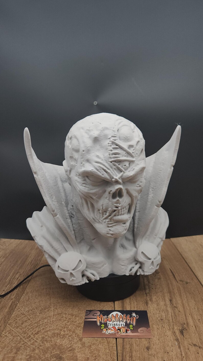 Puede incluir: Escultura detallada en gris claro de una cara esquel&eacute;tica con orejas grandes y puntiagudas y una capa. La escultura tiene una superficie texturizada y est&aacute; montada sobre una base negra. Dos detalles en forma de calavera en la parte delantera.