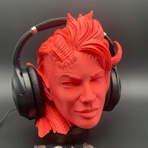 May include: A red headphone stand in the shape of a human head with horns and flowing hair. Black headphones are placed on the stand. The stand has a detailed design and is made of a solid material. The background is dark.