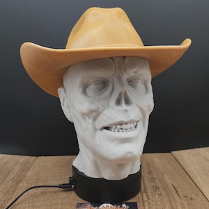 May include: A white, sculpted head of a zombie wearing a tan cowboy hat. The head is mounted on a black base with a power cord. A business card with the text "My Rabbit Trinkets" is in front.