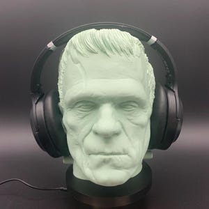 Könnte beinhalten: Eine mintgrüne Frankenstein-Kopfskulptur mit schwarzen Kopfhörern. Die Skulptur hat detaillierte Gesichtszüge und eine Narbe. Die Kopfhörer sind schwarz und vom Over-Ear-Typ. Der Sockel ist schwarz.