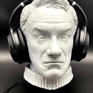 May include: A grey bust of a man with black headphones. The bust has a detailed face and a high, ruffled collar. The headphones are positioned over the ears, suggesting a headphone stand. The bust is made of a textured material.