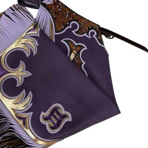 Puede incluir: Chaps de cuero morado con adornos y flecos decorativos dorados y blancos. Los chaps presentan adornos de volutas y un emblema decorativo. Se ve una correa de cuero marrón.