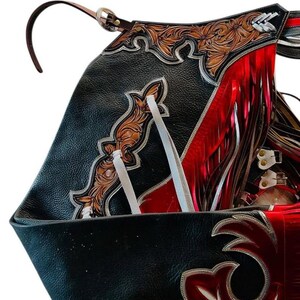 May include: Close-up of a black leather saddle with intricate tooling and red fringe. The saddle features brown floral designs, silver accents, and white ties. A brown leather strap with a silver buckle is visible.