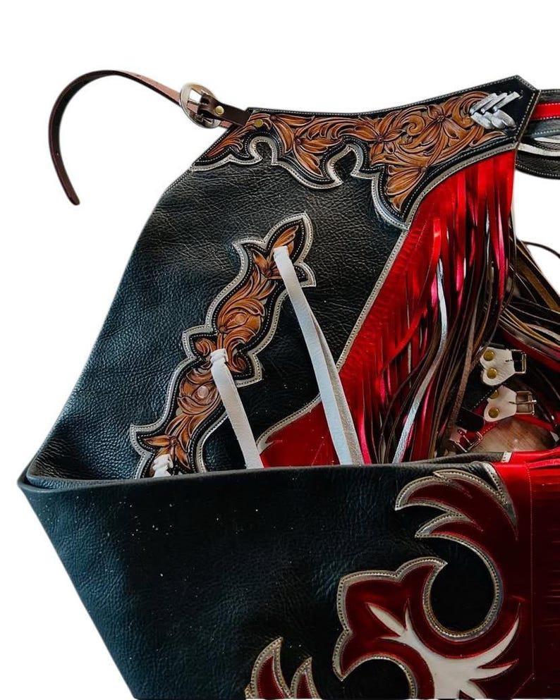 May include: Close-up of a black leather saddle with intricate tooling and red fringe. The saddle features brown floral designs, silver accents, and white ties. A brown leather strap with a silver buckle is visible.