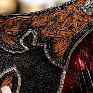 May include: Close-up of a handcrafted leather item with intricate floral tooling. The design features embossed brown flowers and leaves against a black background, accented with silver-toned metal trim and a red, textured section.