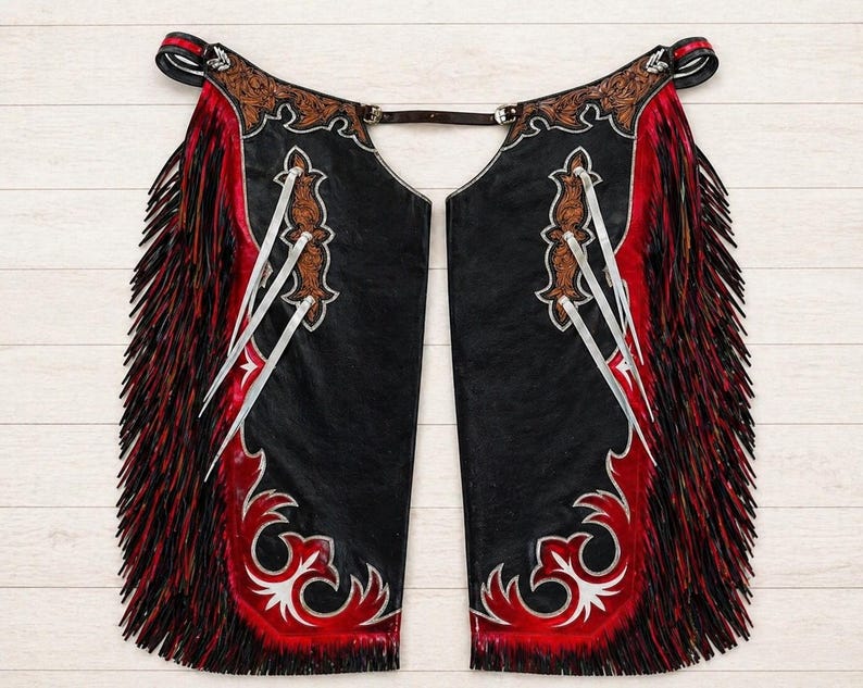 May include: Black leather chaps with red and white decorative accents. The chaps feature a red fringe along the sides and decorative silver-colored embellishments. The top portion has brown leather detailing.