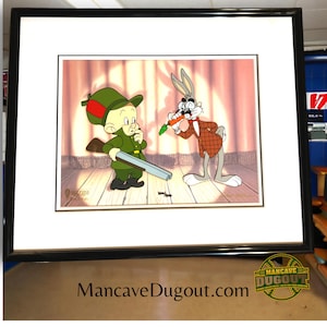 Puede incluir: Celda de dibujos animados enmarcada que presenta a Elmer Fudd apuntando con una escopeta a Bugs Bunny, que está comiendo una zanahoria. La obra de arte está ambientada en un telón de fondo de escenario con un sello de derechos de autor y un número de edición limitada. El marco es negro.