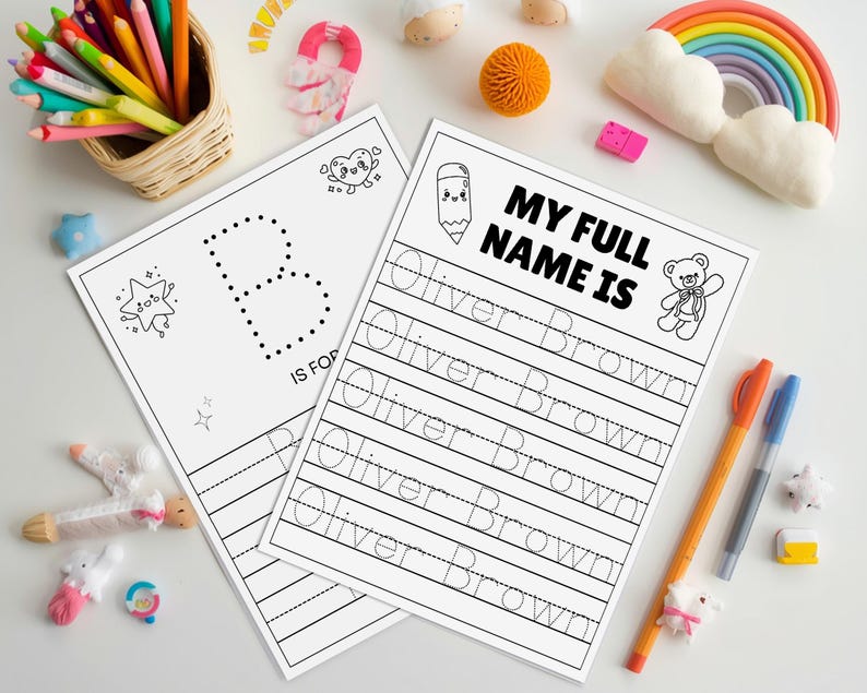 May include: Two white worksheets with black text and illustrations for learning. One sheet says "B IS FOR" with a dotted "B". The other says "MY FULL NAME IS" and includes the name "Oliver Brown" for tracing. Colour pencils and pens are nearby.